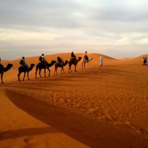 A group traveling on camels over reddish sands under a cloudy sky in a vast desert landscape.